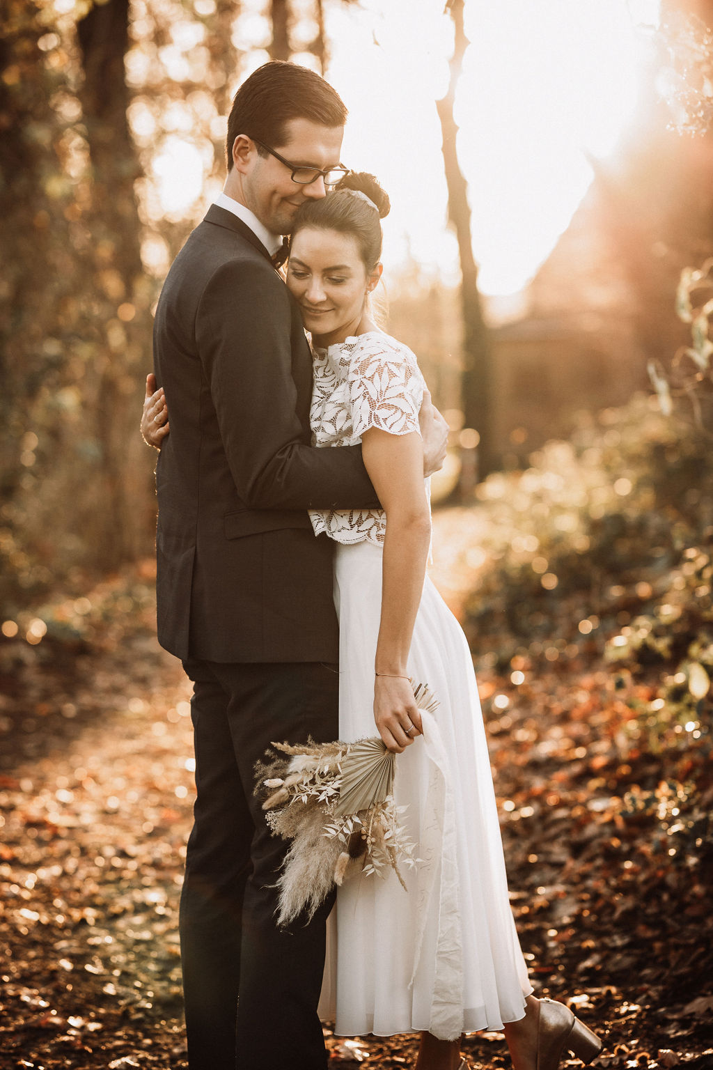 noni Brautmode, Hochzeitsshooting im Wald, Brautpaar händchenhaltend mit Sonneneinfall im Hintergrund. Bräutigam mit dunklem Anzug, Braut in Kurzarm-Kleid mit Boho-Spitze, sich umarmend
