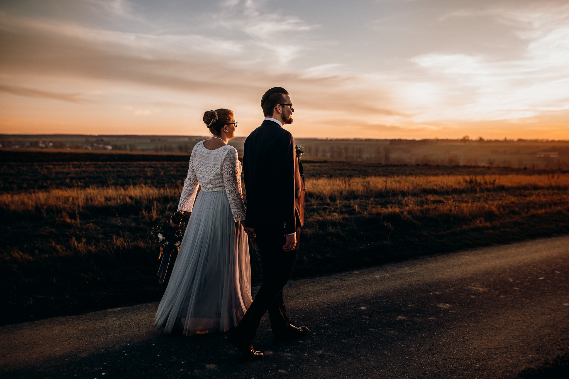 noni Brautmode, Brautpaar bei Sonnenuntergang auf Feldweg, händchenhaltend