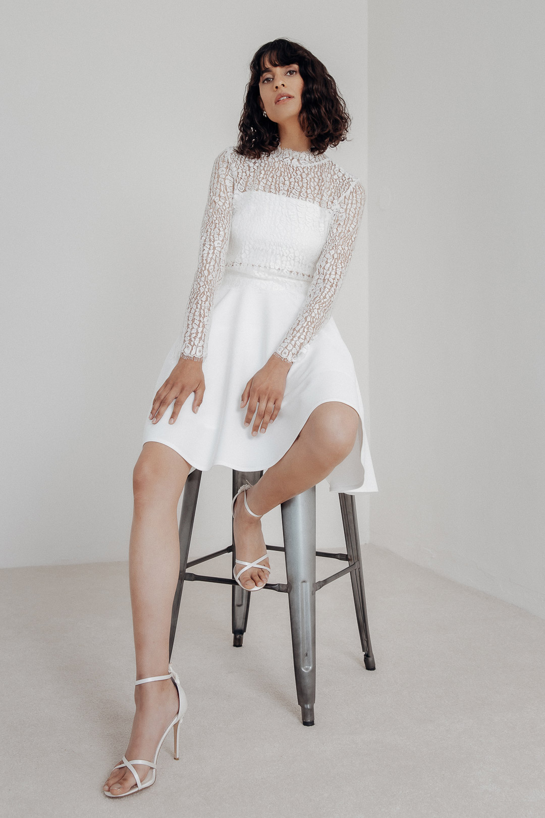 Emina – Stehkragentop aus elastischer Spitze mit Kalea-Rock, brünettes Model im hellen Fotostudio, auf Stuhl sitzend