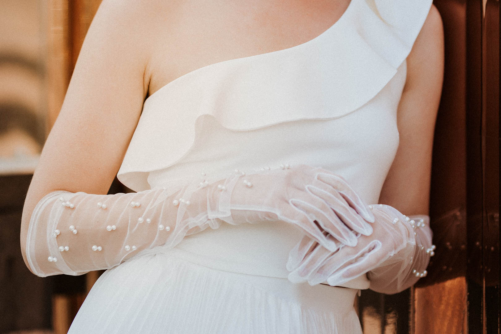 noni Brautmode, Grecia – One-Shoulder Braut Top mit Julica Plisseerock, Outdoor-Aufnahme, blondes Model, Nahaufnahme mit Top und Perlenhandschuhen