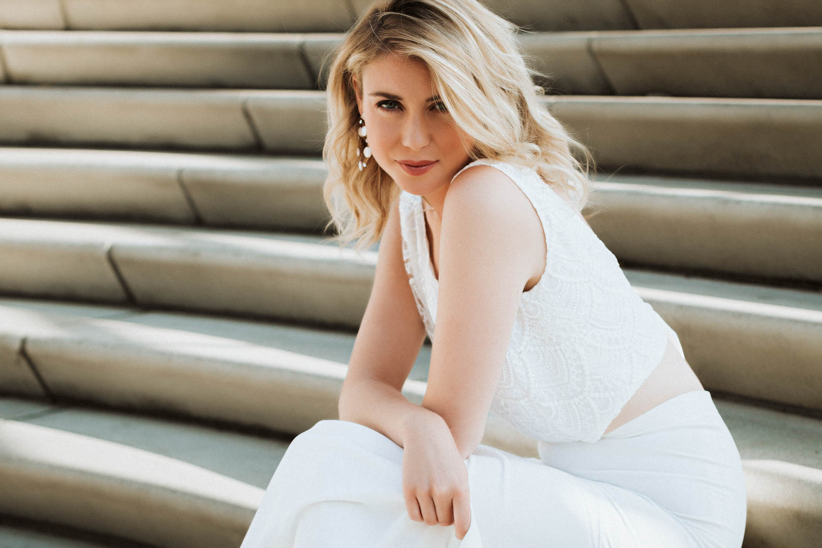 noni Brautmode, Philia – Rückenfreies Braut Top mit Yalena Maxi-Rock, Outdoor-Aufnahme, blondes Model auf Treppe sitzend