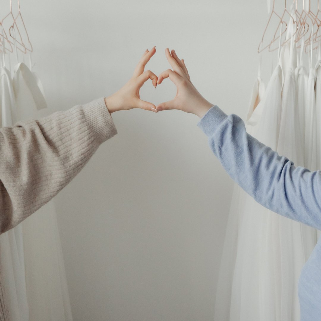 Zwei Hände, die ein Herz formen. Im Hintergrund weiße Brautröcke an Kleiderstange